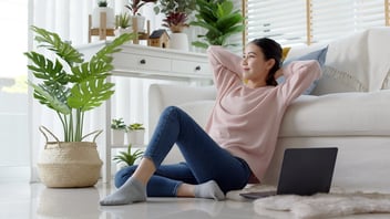 Young attractive beautiful asia female girl or university student sit smile look outside window put arm hand back behind head at sofa couch living room feeling relax comfort at cozy home houseplant.